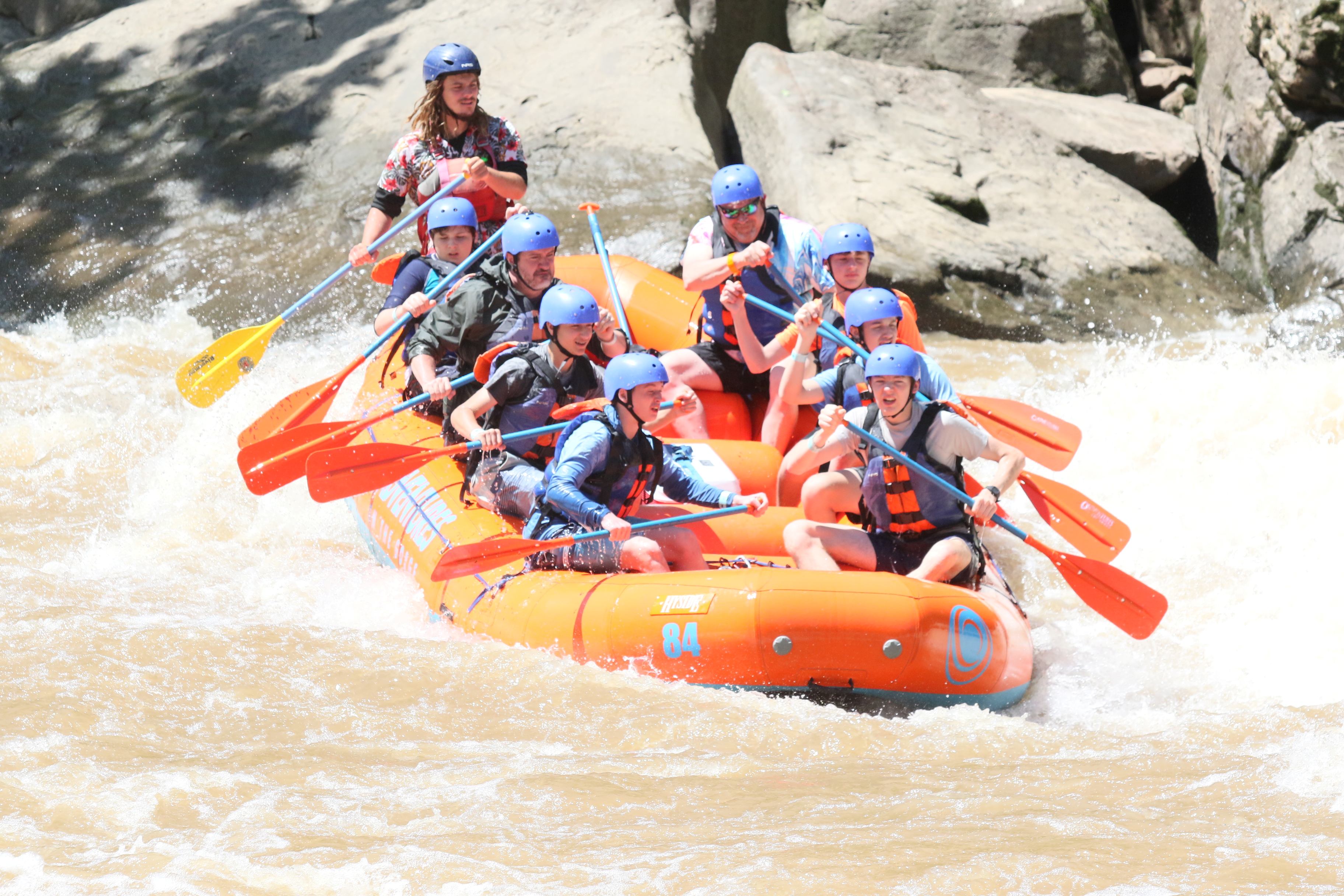 Scouts and adult advisors paddling a raft on the New River through fast-moving water and rocky riverbanks, surrounded by steep wooded hills and a sense of adventure and teamwork.